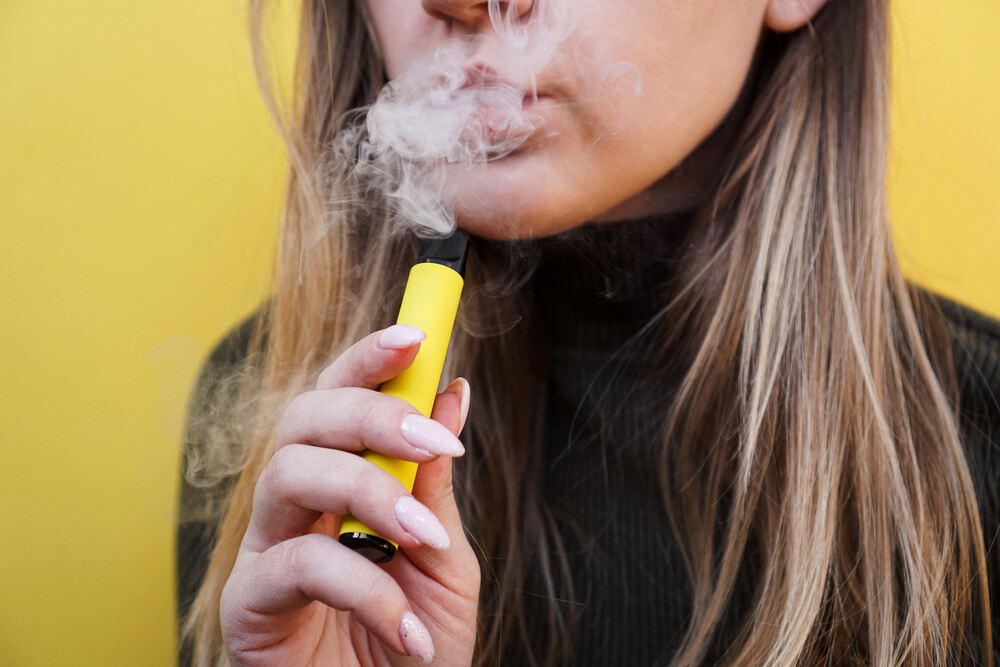 A young woman smokes a Delta 8 vape in front of a yellow background.