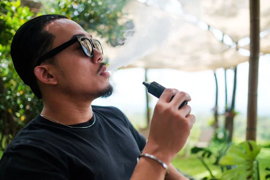 A man wearing sunglasses vapes outdoors.
