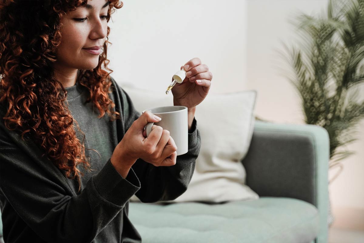A woman holds a cup of coffee, adding drops of CBD oil to it.