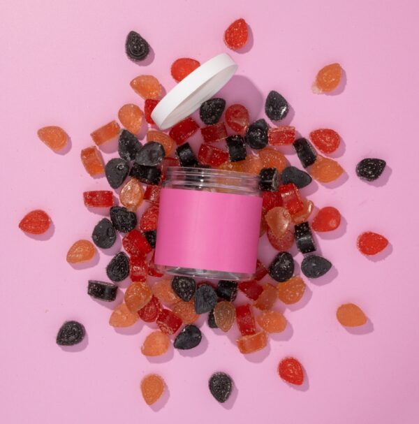 A jar of CBD gummies sits open with the gummies spilled on a pink background.