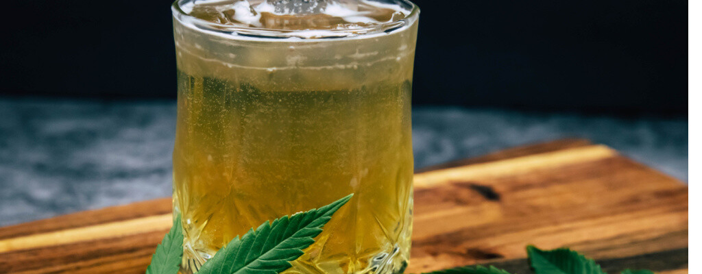 Hemp leaves sit next to a cold, amber-colored drink in a short glass.