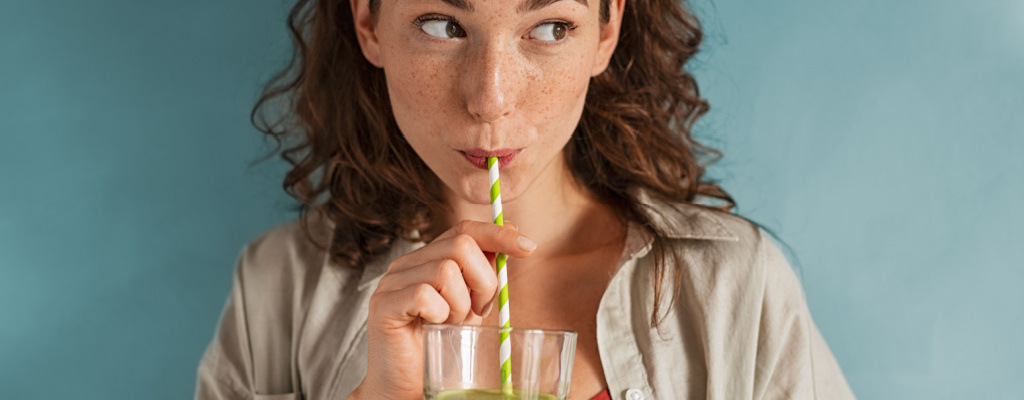 A young woman drinks a green smoothie in front of a blue background.