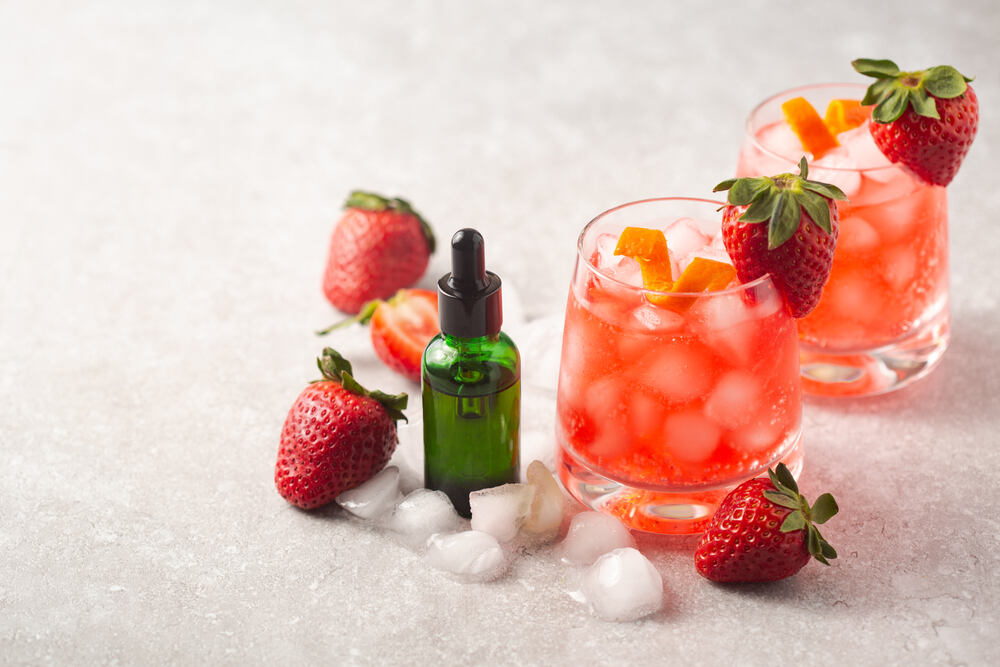 Strawberry-flavored THC drinks, ice cubes, strawberries, and a bottle of tincture sit on a table. 