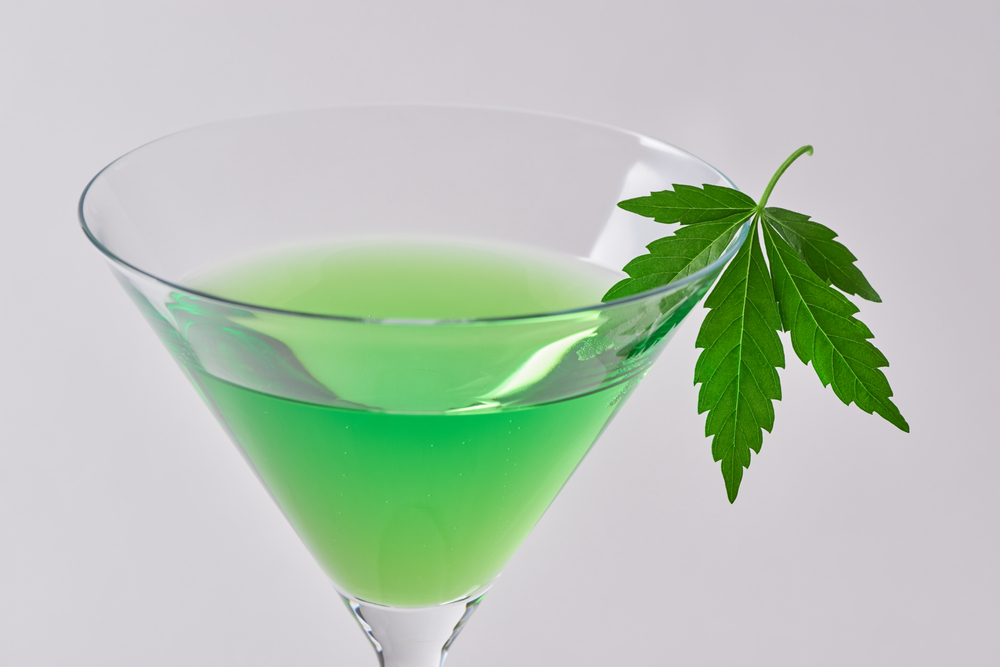 A hemp leaf hangs on the edge of a martini glass containing a green cannabinoid drink.