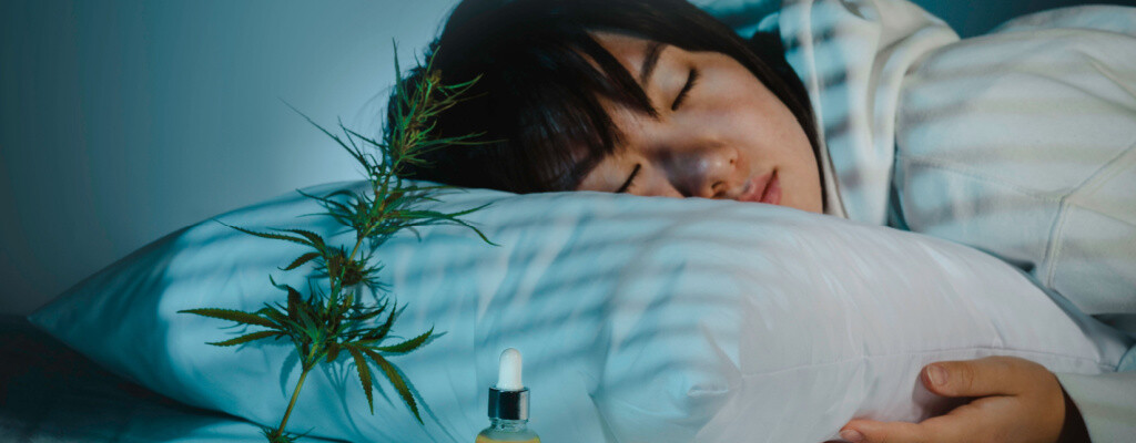CBD oil and a hemp leaf sit in front of a woman sleeping soundly on a bed.