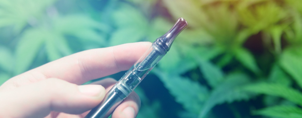 A hand holds a vape pen in front of a background of hemp leaves.