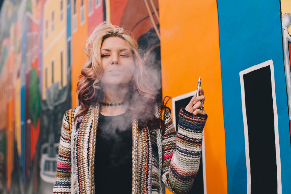 A woman uses a vape in front of a large mural. 