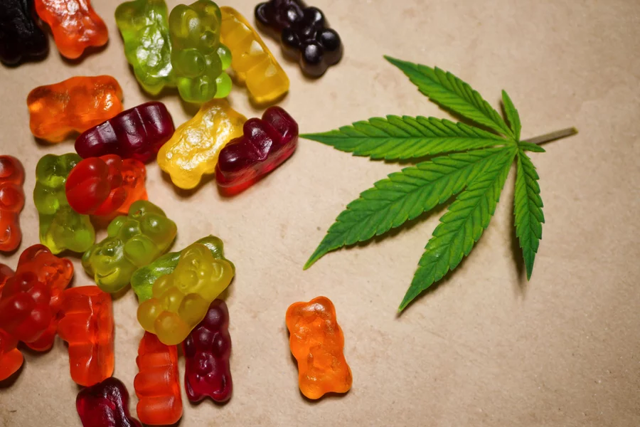 Multicolored gummies sit next to a hemp leaf on a table in an overhead view.