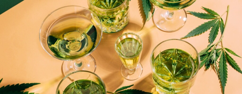 Hemp leaves and cocktail glasses filled with green liquid rest on a table. 