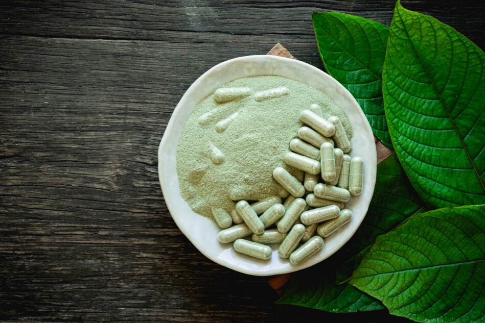 Kratom powder, capsules, and fresh leaves shown sitting on a table from above.