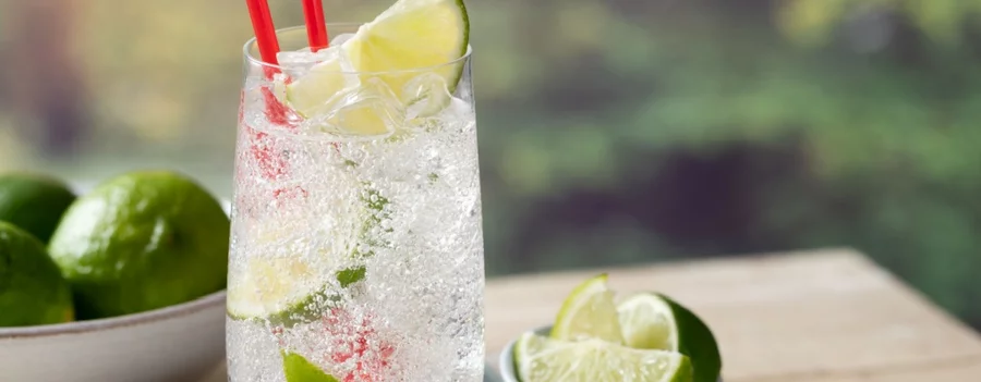 A kratom seltzer in a tall glass with ice, lime wedges, and a straw sits on a table.